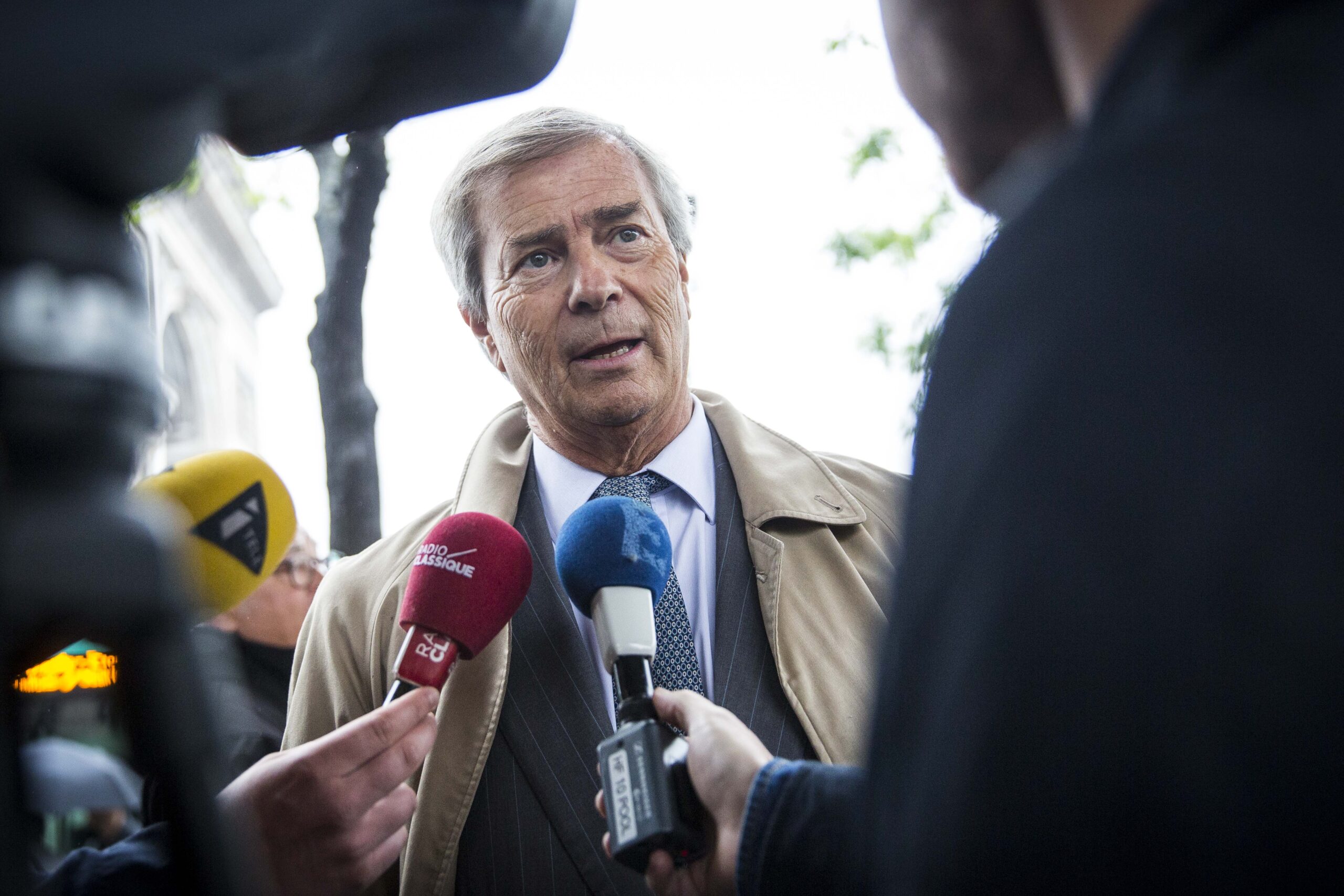 Bollore, in a tan lapel trench coat over a suit and tie, speaks to members of the press. Two microphones are in front of him from French media.