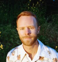 Miles is seated in front of a green garden bush, wearing a white collared shirt with red/orange flowers 